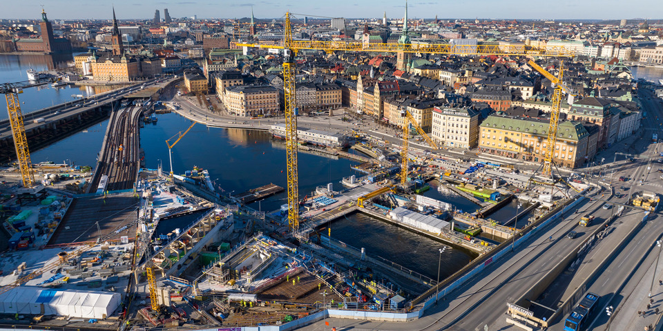 Drönarvy över arbetsområdet vid Slussen. På vattnet mellan Södermalm och Gamla stan pågår byggarbeten. Inne på arbetsområdet finns fem gula lyftkranar.