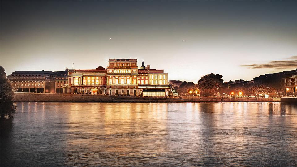 Visualisering av Kungliga Operan i Stockholm med en föreslagen påbyggnad, kvällstid sett från Slottskajen. Byggnaden är upplyst och speglas i vattnet.