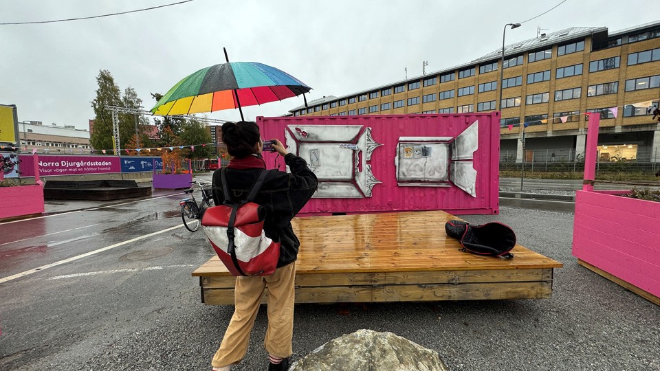En person med paraply fotograferar en rosa container med silvrigt konstverk. 