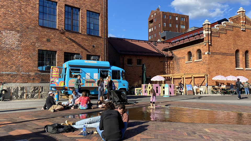 Torg med människor som sitter på marken. Tegelbyggnader omringar torget. 