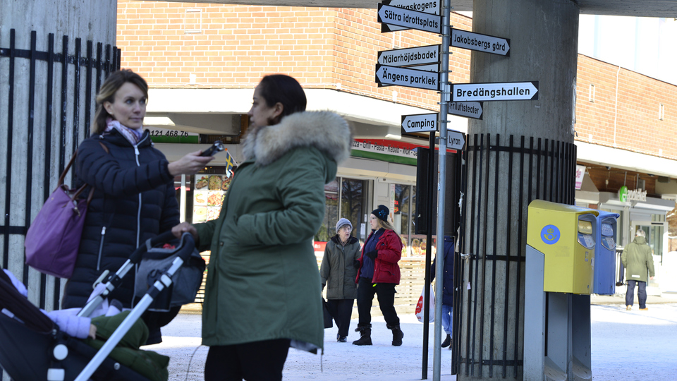 En kvinna intervjuar en kvinna med barnvagn i ett centrum. Tegelbyggnad, folk och vägskyltar i bakgrunden.