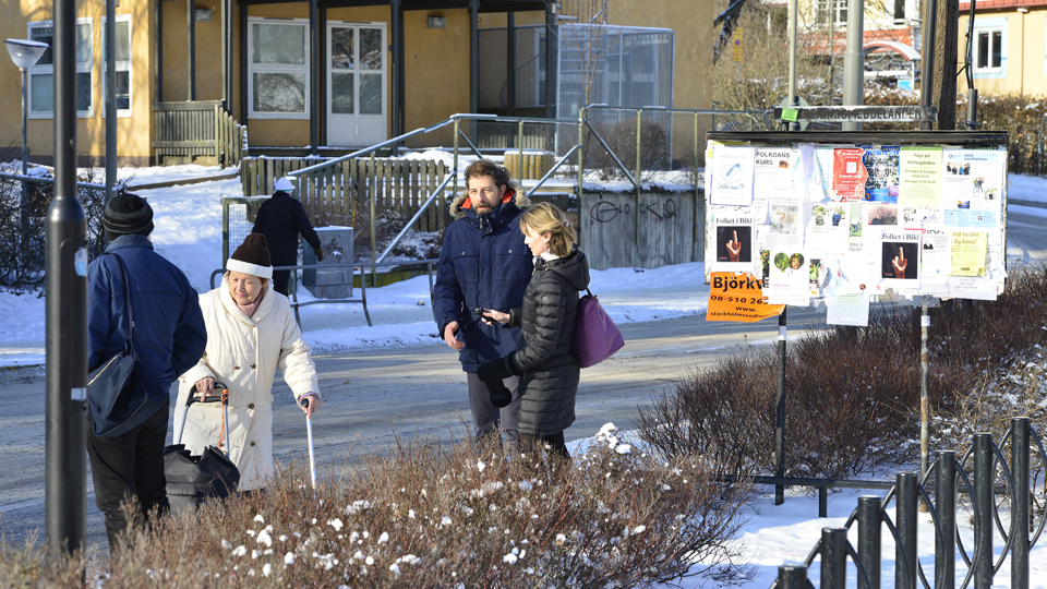 Några personer intill en anslagstavla. En gul byggnad och snöiga trottoarer i bakgrunden.