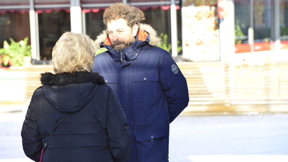 En man intervjuar en kvinna i vinterjacka på ett solbelyst torg. Hon syns bakifrån.