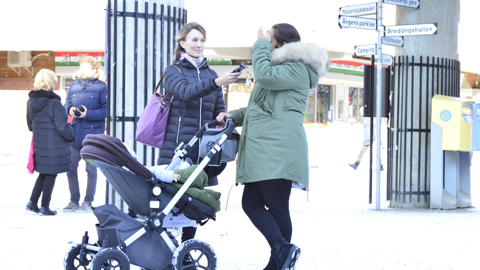 En kvinna intervjuar en kvinna med barnvagn i ett centrum. Tegelbyggnad, folk och vägskyltar i bakgrunden.