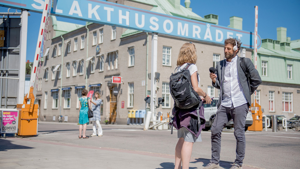 En man med mikrofon intervjuar en kvinna framför en vägbom med skylten Slakthusområdet.