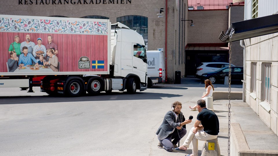 En man intervjuar en man som sitter på en betongsugga. I bakgrunden syns en lastbil.