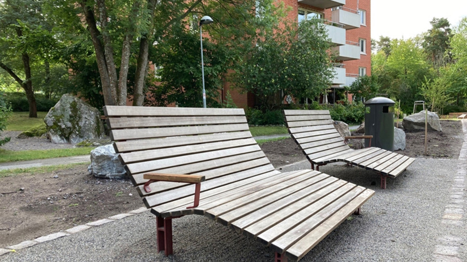 Liggande träbänkar vid grusplan, hus i tegel, gångväg och träd i bakgrunden.