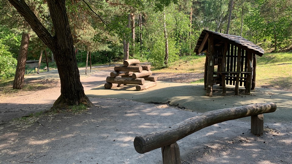 En lekplats i skogen med ett litet lekhus och klätterredskap av stockar. 