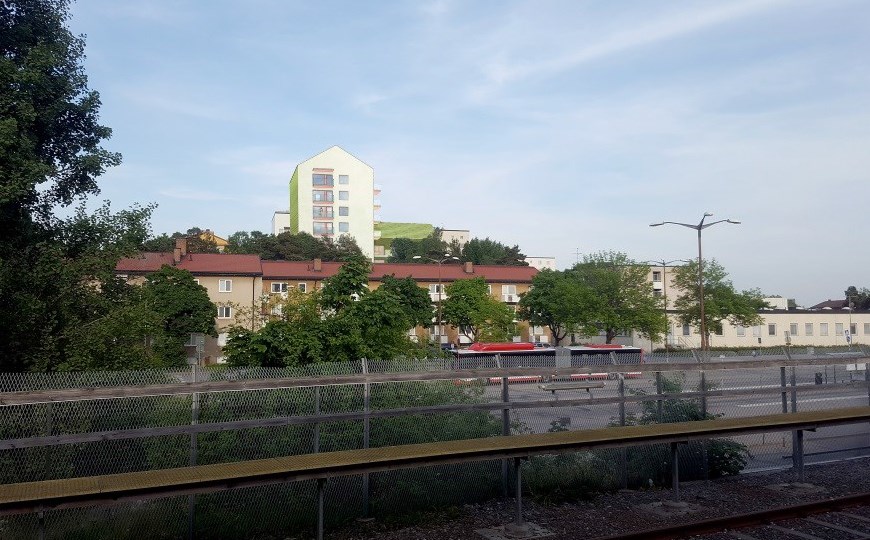 Vy från perrong och spårområde, i bakgrunden syns ett flerbostadshus bakom lägre hus, illustration.