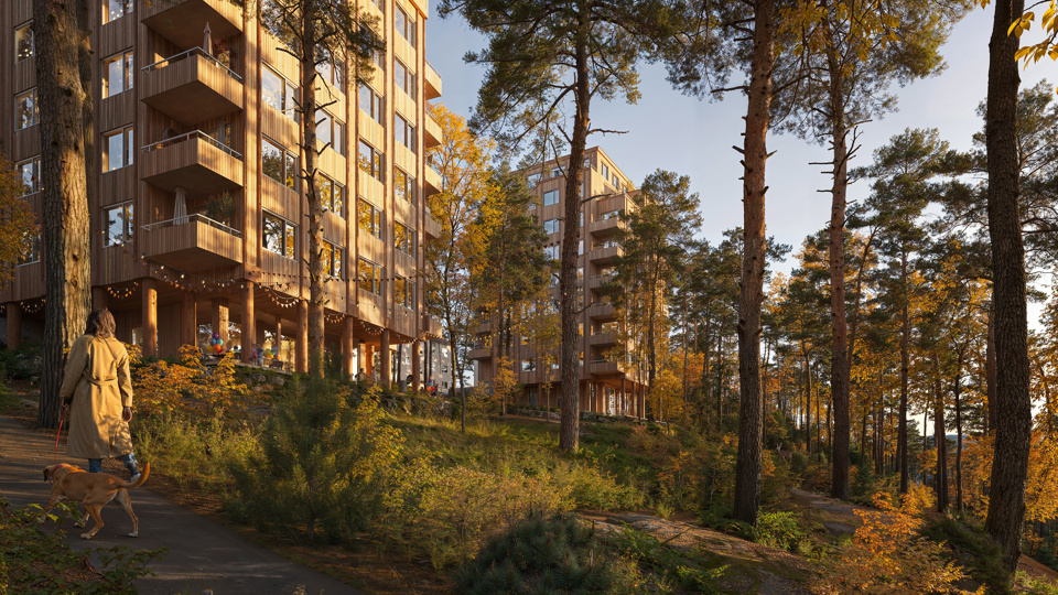 Visionsbild som visar den nya bebyggelsen sett från gångstråket från Gröndalsvägen till Fregattvägen. De två västligaste husen syns. Byggnaderna är gjorda i trä med indragna balkonger i hörnen. Längst ner i mötet med naturen syns en indragen suterrängvåning och pelare som grundlägger byggnaden ovanför. I förgrunden visas naturmark med berg och växtlighet samt flera tallar. Även mellan de två husen finns träd. I det vänstra hörnet visas gångstråket med en person som går med en hund.   