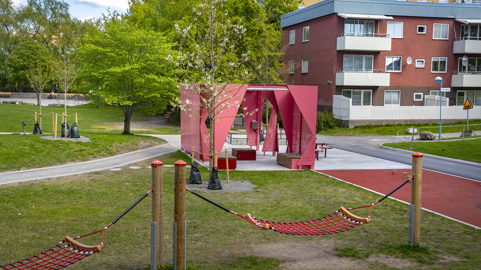 Hängmattor på en gräsyta, framför en paviljong i röd plåt med sittplatser.