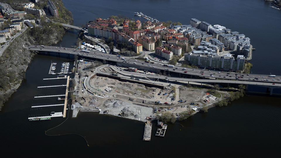 Flygvy över området, en ö, med en motorvägsbro som går rakt över. På ena sidan motorvägen syns flerbostadshus och på andra sidan är byggarbetsplats. Båtbryggor, vatten, fotografi.