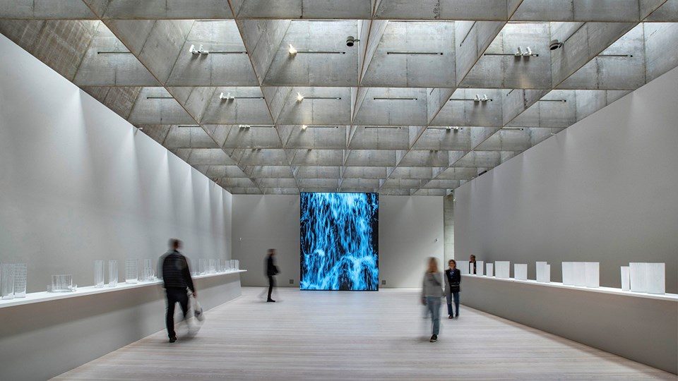 Utställningslokal med ljusinsläpp från taklanterninerna och en digitalskärm med ett konstverk, foto.