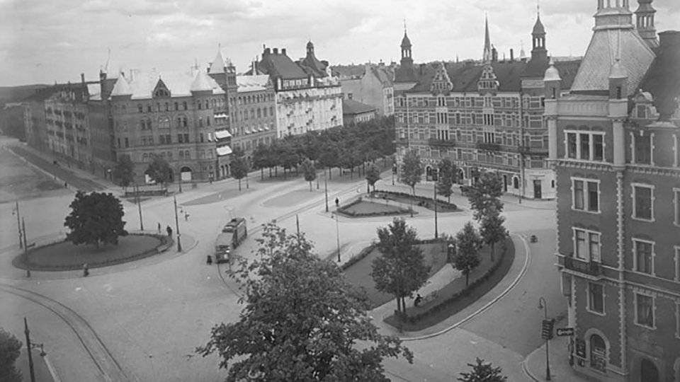 Stenbelagt öppet torg med spår för spårvagn, omgivet av hus. Gröna ytor och träd, svartvitt foto.