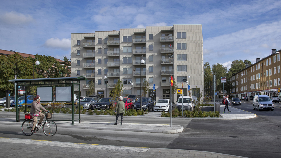 Busshållplats, person på cykel, människor i rörelse. I bakgrunden syns flerbostadshus och parkeringar, fotografi.