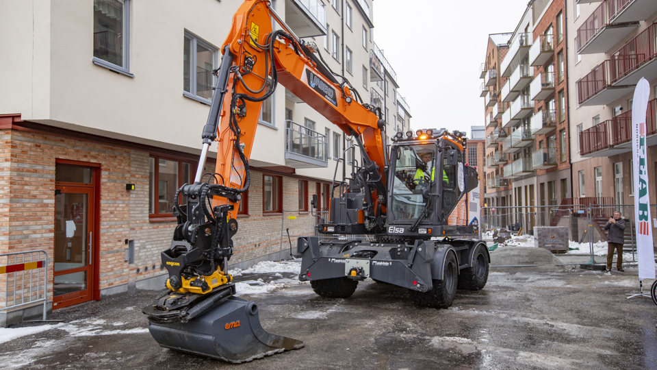 En elgrävare parkerad intill ett nybyggt flerbostadshus. Skopan är nedfälld mot marken.