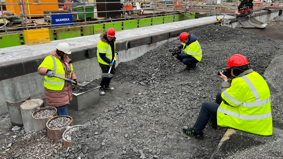 Två personer med gula västar och bygghjälmar står inne på byggarbetsplats, på varsin sida om en låda av plåt. Personerna håller i varsin spade och lägger grus ovanpå lådan. Framför de två personerna och lådan sitter två personer, också iklädda gula västar och bygghjälmar. De fotar händelsen.