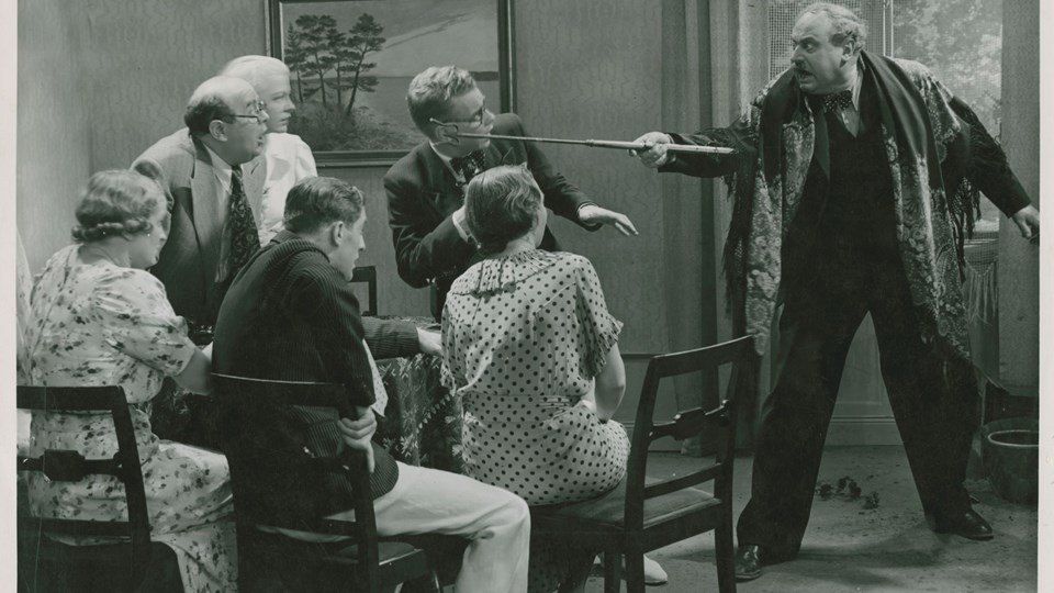 Svartvit bild från 1930-talet. en stor man med en sjal över axlarna pekar med en eldgaffel mot en grupp av tre kvinnor och tre män.
