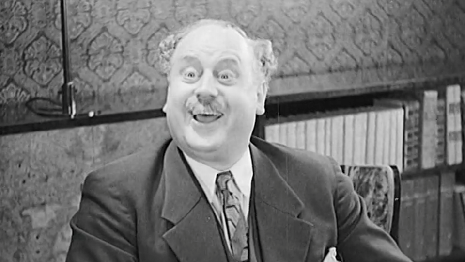 Svartvit bild från 1930-tal. En stor man med mustasch och kostym sitter vid ett bord. Han ser glad och förvånad ut.