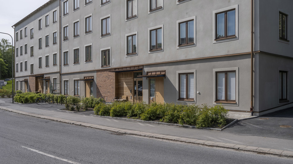 Grått flerbostadshus med bruna fönsterkarmar som ligger intill bilväg, foto. 
