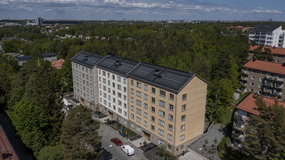 Översiktsbild på flerbostadslänga med solceller på taket. Huset är omgivet av trädtoppar, foto.