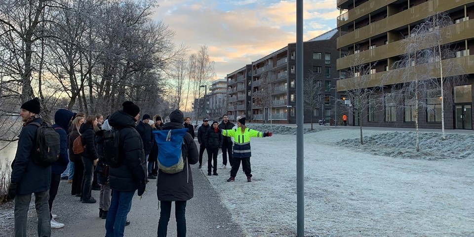 En grupp människor som står på en gräsmatta. Hus till höger i bild. 