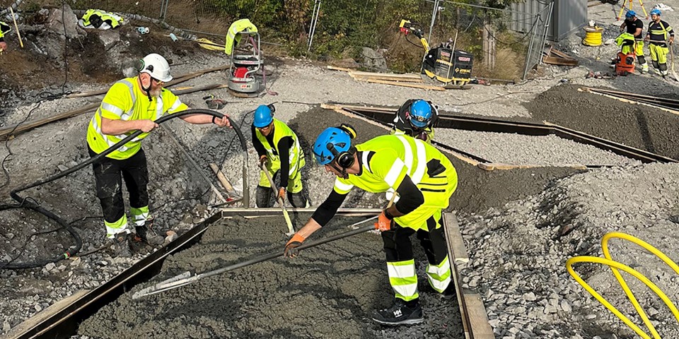 Tre byggarbetare i gula kläder häller cement i en stor form i en backe. 