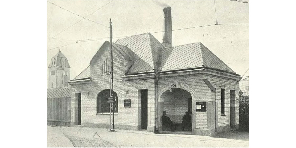 En historisk svartvit bild av en liten tegelbyggnad med valvbågar, tagen år 1914. Byggnaden har ett brant tak med en skorsten, och två personer sitter under en täckt ingång.