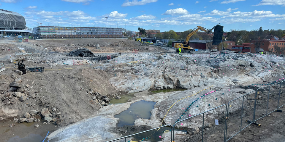 En byggarbetsplats i Slakthusområdet där den andra och sista etappen av marksänkning pågår. Maskiner och utrustning används för att gräva och schakta bort massor. I bakgrunden syns byggnader och en stor arena under klarblå himmel med lite moln.