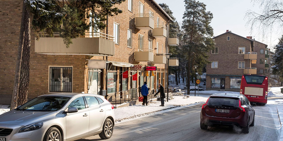 Bostadshus i tegel med frisörverksamhet på markplan. Parkerade bilar, omgivande träd och människor som samtalar. Foto.