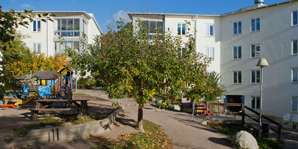 Innergård med lekplats, träd och bänk med bord. Omgiven av fler bostadshus, foto.