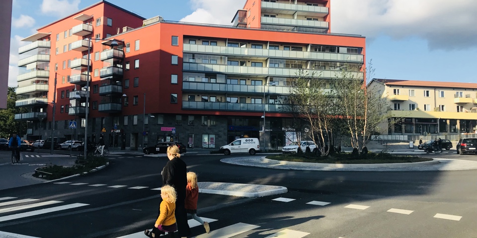 Bostadshus i olika nivåer med verksamhetslokaler på bottenplan, bakom en rondell. Människor i rörelse, fotografi.