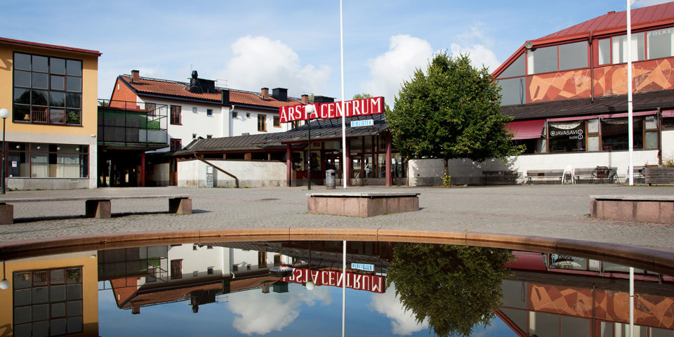 Ett stenbelagt torg med med en damm och sittplatser. Omslutet av hus, bibliotek och kafé, foto. 