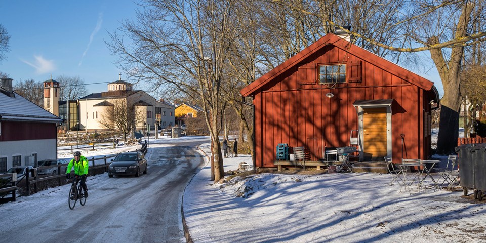 Litet rött gårdshus med cafébord och stolar framför, på vägen cyklar en person. I bakgrunden syns en kyrka, fotografi.