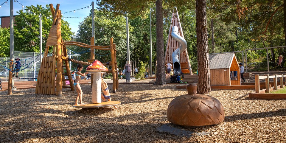 Bandängens lekplats med gungor, rutschkana och klätterställning, omgiven  träd av.  Barn som leker, foto.