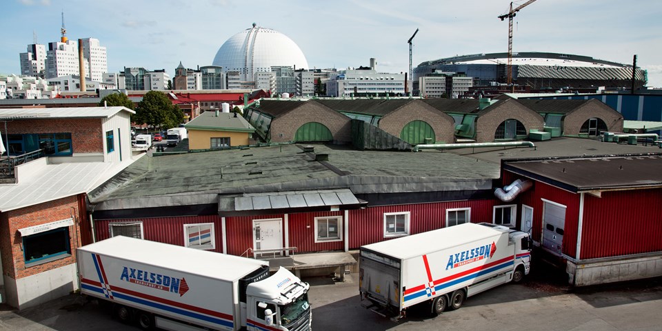 Varuintag på baksidan av låg byggnad. Två lastbilar. I bakgrunden flera byggnader samt Globen och Tele 2 Arena.  Foto.