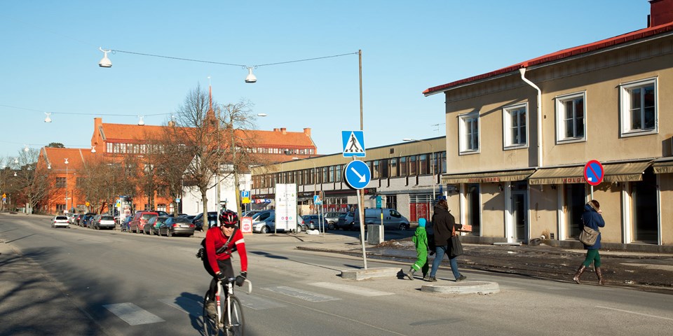 En bilväg genom bostadsområde med övergångsställe. En cyklist och människor som korsar vägen, foto.