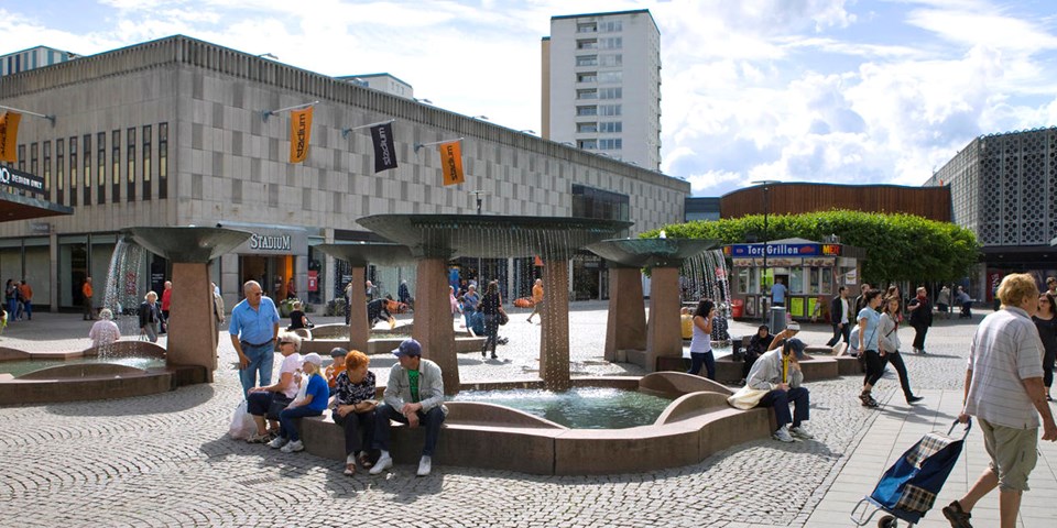 En torgyta med fyra fontäner omgiven av butiker. På torget finns en grillkiosk  och människor i rörelse. Foto.