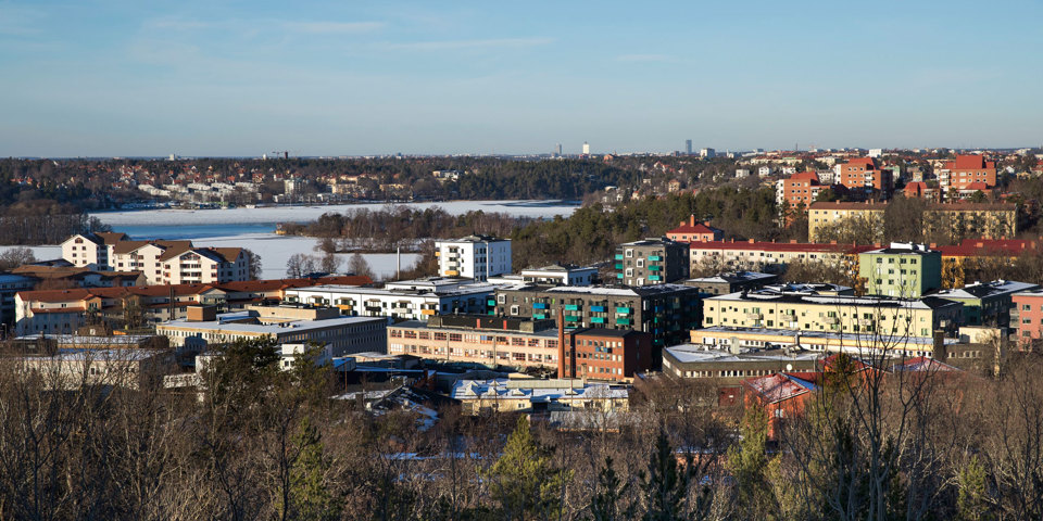 Vy över Hägersten. I bakgrunden vatten, i förgrunden träd. Fastigheter för bostäder och olika verksamheter. Foto.