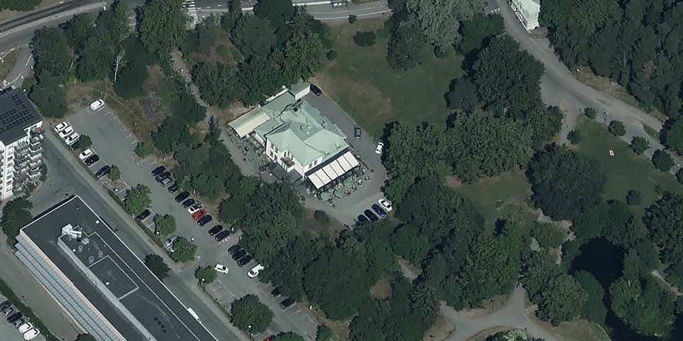 Flygvy över området, byggnad mitt i bild med ljusgrönt tak, parkering, skogsmiljö, fotografi.