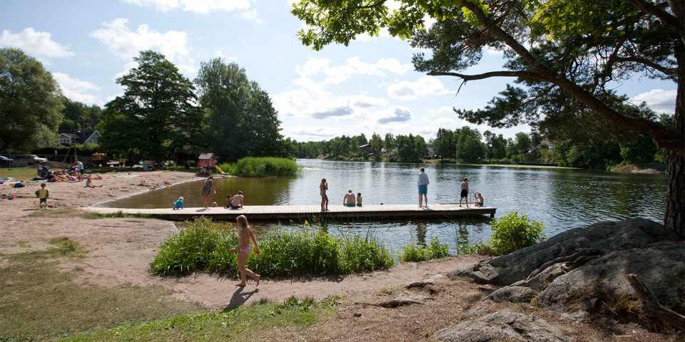 Badplats med sandstrand, gräsmatta, brygga, träd och små klippor. Människor som badar och solar. Foto.