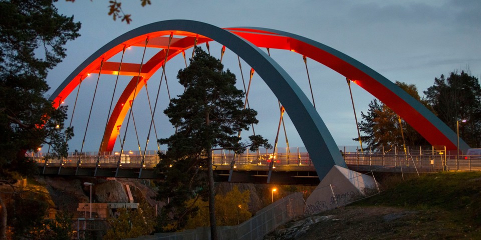 En upplyst bro i skymningen. Tallar på båda sidor av bron, berg i förgrunden, foto.