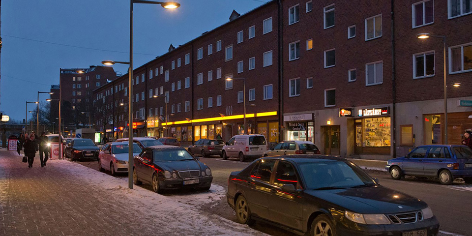 En gata som är upplyst av lyktstolpar. Parkerade bilar på gatan. Upplysta affärer. Människor i rörelse. Foto.