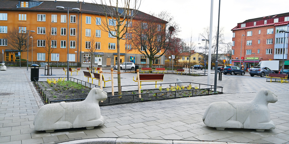 Torg med bänkar och träd omslutet av färgglada flerbostadshus, foto.