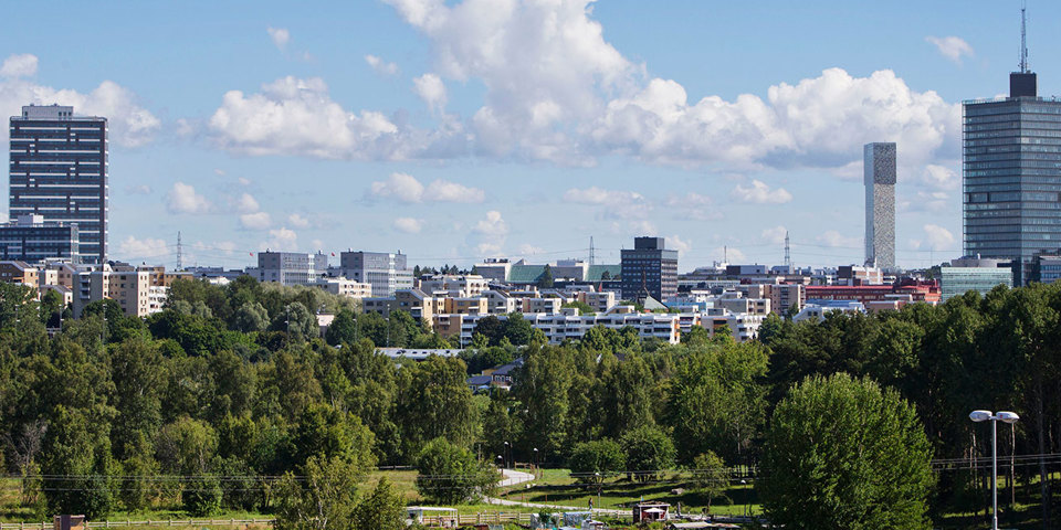 Grönområde med träd och buskar.  Ett stort antal byggnader i olika storlekar i bakgrunden. Foto.