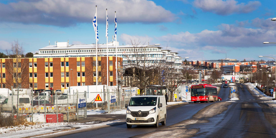 En bilväg med körande bilar. Invid vägen finns industribyggnader och parkering med lastbilar, foto.