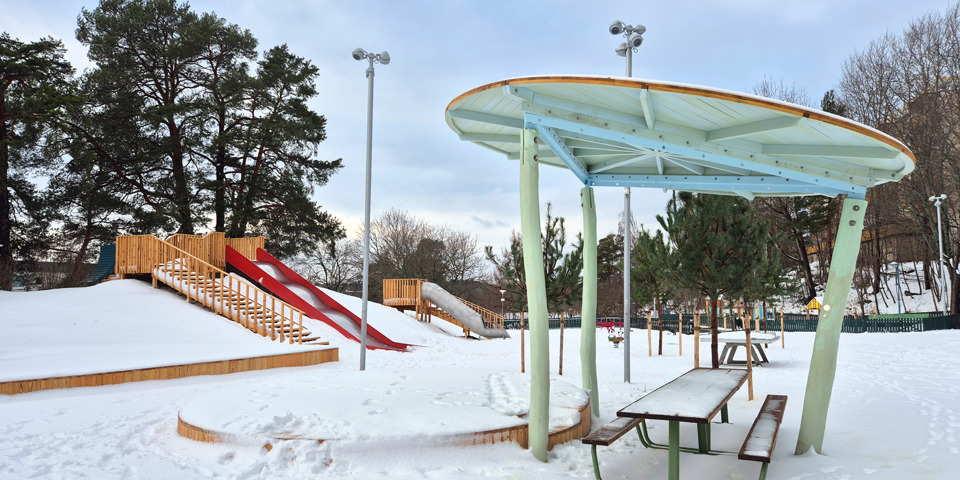 En snötäckt parklek. En parkbänk med tak över syns framför en backe med rutschkanor.