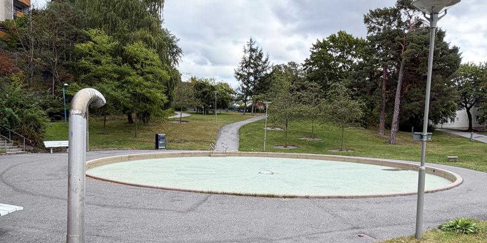 Asfalterad yta med cirkelformad plaskdamm i mitten. Bostadshus, gångväg och träd som omger.