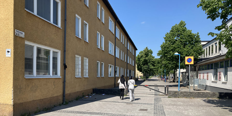 Gångstråket Rinkebygången omgiven av byggnader. Två personer promenerar. Längre fram längs gatan syns träd