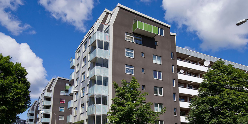 Bostadshus som är flera våningar högt. Huset har balkonger och loftgångar. Träd och lyktstolpar utanför huset. Foto.